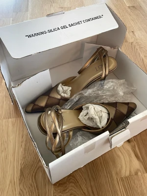 Bruna pumps med rutmönster och remmar - Helt nya Snygga pumps från KIT med spetsig tå och rutmönster i bruna och beige nyanser. Skorna har tunna, korsade remmar i metallic och en elegant klack.  lyxig känsla och passar perfekt till trendiga outfits. Storlek 38!