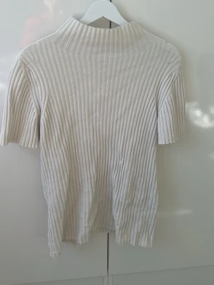 Ribbad vit polotopp - Säljer en ribbad vit polotopp med korta ärmar. Toppen har en hög krage och är tillverkad i ett mjukt, stretchigt material som sitter snyggt på kroppen. Perfekt att styla med jeans eller kjol för en clean look.