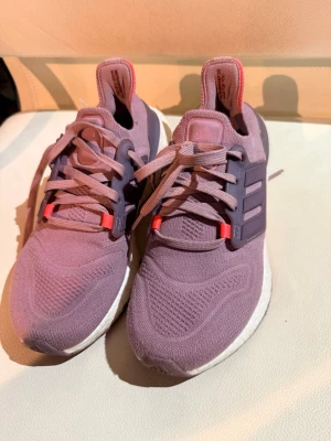 Lila Adidas Ultraboost sneakers - Säljer ett par lila Adidas Ultraboost sneakers med mesh-ovandel och vita Boost-sulor. Skorna har lila snören, mörklila detaljer och röda accenter vid snörningen. Perfekta för löpning eller gym och riktigt sköna på foten.