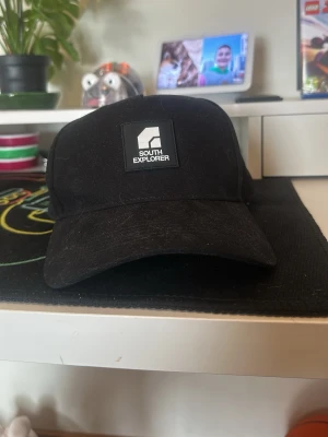 Svart keps från South Explorer - Snygg svart keps från South Explorer med böjd skärm och justerbar snapback baktill. Kepsen har en stilren patch med logga framtill och är tillverkad i ett slitstarkt tyg som passar perfekt till streetwear-looken. Är lite hårig men tvättas vid köp, använder inte för jag har andra kepsar hemma. 