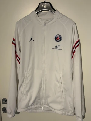 Vit PSG träningsjacka Jordan - Säljer en vit träningsjacka från Jordan x PSG med dragkedja, två fickor och klubbmärke på bröstet. Jackan har röda och blå ränder på ärmarna och elastiska muddar. Perfekt för dig som gillar fotboll och streetwear.