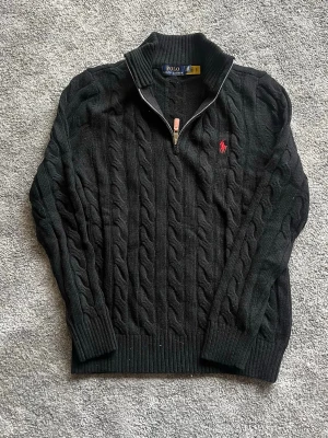 Polo Ralph Lauren Black Cable Knit 1/4 Zip Jumper - Classic Polo Ralph Lauren quarter-zip sweater in a timeless black cable knit. Features the iconic red embroidered pony logo on the chest and a leather zipper pull detail. • Condition: Great pre-owned condition. Well-maintained with plenty of life left. • Fit: Slim/Regular fit (check your specific tag for the size, looks like a Men's size). • Style: 1/4 zip mock neck, perfect for layering over a shirt or wearing casually. • Material: 100% Cotton (heavyweight and high quality). 