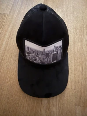 Svart truckerkeps från Gårda - Snygg svart truckerkeps från Gårda med mesh på baksidan och en stor svartvit New York-print framtill. Kepsen har böjd skärm och justerbar passform. Perfekt för dig som gillar streetstyle och vill ha en unik accessoar.