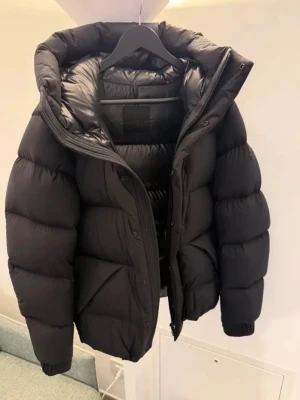 Moncler madeira  - Snygg svart dunjacka från Moncler med stor huva och quiltad design. Jackan har dragkedja och tryckknappar framtill samt elastiska muddar vid ärmsluten. Perfekt för kalla vinterdagar och riktigt stilren look.