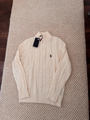 Stickad krämvit tröja Polo Ralph Lauren - Snygg stickad tröja från Polo Ralph Lauren i krämvit färg med klassiskt flätmönster. Tröjan har en hög ribbad krage med dragkedja och den ikoniska logon broderad på bröstet. Perfekt för att hålla sig varm och stilren under kyliga dagar.