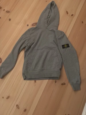 Grå hoodie från Stone Island - Snygg grå hoodie från Stone Island med klassisk patch på vänster ärm. Hoodien har en stor ficka framtill och ribbade muddar vid ärmslut och nederkant. Perfekt för en avslappnad och trendig stil. Tillverkad i mjukt bomullsmaterial. Passar dig som är ungefär 160 cm
