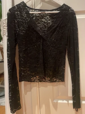 Svart spetsblus från Gina Tricot - Snygg svart långärmad topp från Gina Tricot i genomskinlig spets med blommönster. Djup v-ringning och figurnära passform, perfekt för att lyfta vilken outfit som helst. Materialet är stretchigt och mjukt för en bekväm känsla.