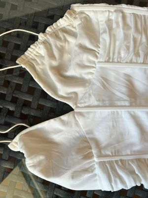 Vit korsettopp med lite volanger  - Trendig vit korsettopp med korta ärmar och smockdetaljer. Toppen har snörning vid axlarna och markerade sömmar framtill för en snygg siluett. Perfekt för dig som vill ha en chic och modern look. Tyget har gått upp lite inuti 