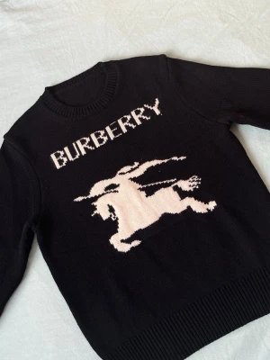 Svart stickad Burberry-tröja - Cool svart stickad tröja från Burberry med ett stort vitt motiv av en riddare och Burberry-loggan på bröstet. Tröjan har rund halsringning, långa ärmar och ribbade muddar. Perfekt för dig som gillar streetstyle och exklusiva märken.