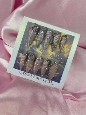 Exlipse (S); Long Stiletto - Unika handgjorda press-on naglar i rosa cateye med kromad finish och silverdetaljer. Naglarna har en spetsig stiletto form och dekoreras med handmålad 3Dmönster och små metalliska pärlor accenter. Perfekt för dig som vill sticka ut med en edgy och kreativ nageldesign.S: 15 mm, 11,5 mm, 12 mm, 11 mm, 9 mm 