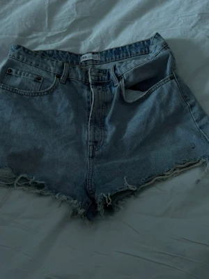 Blå denimshorts från Zara - Säljer ett par klassiska blå denimshorts från Zara med råa, fransiga kanter och femficksmodell. Shortsen har hög midja och en avslappnad passform, perfekta för varma dagar. Stängs med knapp och dragkedja framtill.