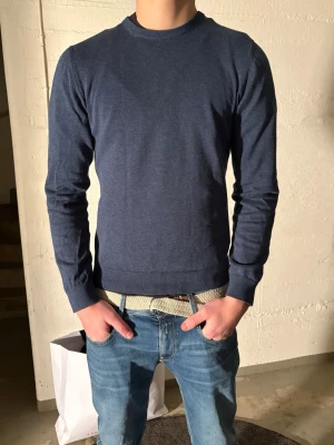 Mörkblå rundhalsad tröja - En stilren mörkblå tröja med rund halsringning och långa ärmar. Tröjan har ribbade muddar vid ärmslut och nederkant, vilket ger en snygg passform. Perfekt att matcha med jeans för en enkel och clean look.