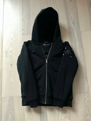 Svart hoodie från Moose Knuckles - Snygg svart hoodie från Moose Knuckles med dragkedja framtill och huva. Har en ikonisk logga i metall på ärmen och mjukt foder med mönster. Perfekt för dig som vill ha en stilren och exklusiv look. Materialet känns tjockt och kvalitativt. Perfekt för året om, hel svart är den🌟