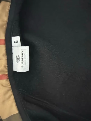 Svart hoodie från Burberry M - Snygg svart hoodie från Burberry i storlek M med klassiskt broderat Burberry-märke på bröstet och ikoniskt beige rutigt foder i huvan. Perfekt för dig som vill ha en stilren och lyxig look. Mjuk bomullsblandning och bekväm passform.