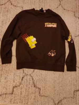 Brun Simpsons sweatshirt med tryck från HM - Cool brun sweatshirt med tryck av Bart Simpson och Itchy & Scratchy på framsidan. Texten 'Bartholomew J. Simpson' syns på bröstet och ärmen har gul text. Tröjan har rund hals och ribbade muddar. Perfekt för dig som gillar tecknade serier och streetwear.