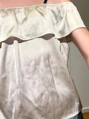 Champagnefärgad axelbandslös topp - Säljer en champagnefärgad axelbandslös topp i glansigt material med volangdetalj upptill. Toppen har en lös passform och är perfekt för dig som gillar en enkel men ändå elegant stil. Passar till jeans eller kjol för en trendig look.