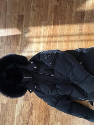 Svart dunjacka med pälskant - Snygg svart dunjacka från Moose Knuckles med stor huva och svart pälskant. Jackan har flera fickor med dragkedja, silverfärgade detaljer och logotyp på ärmen. Perfekt för kalla vinterdagar och riktigt varm. Står XXL men passar mindre XL större L