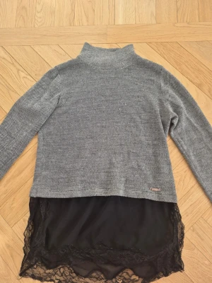 Grå stickad tröja med spets från Guess - Snygg grå stickad tröja från Guess med hög krage och långa ärmar. Tröjan har en unik detalj med svart spetskant och tyg i nederdelen som ger en lager-på-lager-look. Perfekt för dig som vill ha något extra till din outfit.