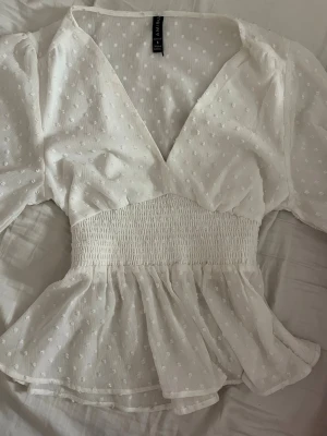 Vit prickig blus - Söt vit blus med diskreta prickar i tyget, v-ringning och smockad midja. Blusen har volang nedtill och långa puffärmar med smock vid ärmsluten. Tillverkad i ett tunt, lätt material som ger en luftig känsla.