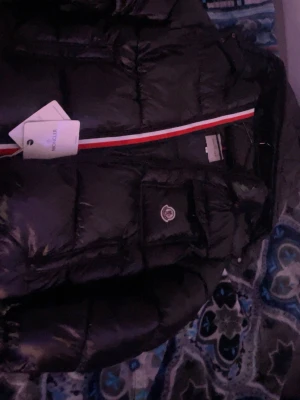 Svart dunjacka från Moncler - Snygg svart dunjacka från Moncler med glansig finish och klassisk Moncler-logga på bröstet. Jackan har en röd, vit och blå randig detalj på insidan och praktisk bröstficka med lock. Perfekt för kalla dagar och riktigt trendig stil.