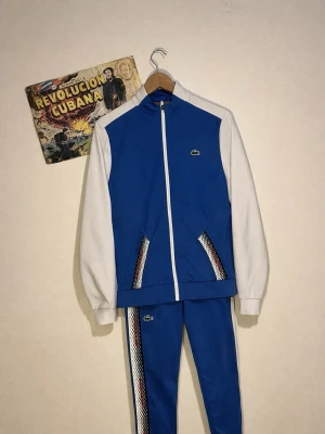 tracksuit Lacoste - Säljer en snygg blå och vit tracksuit från Lacoste. Jackan har vita ärmar, blå kropp och dragkedja framtill samt coola mönstrade detaljer vid fickorna. Byxorna är blå med matchande mönstrad rand längs benet och Lacoste-logga på både jacka och byxa.