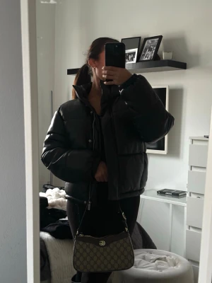 Svart pufferjacka med hög krage - Säljer en svart pufferjacka med hög krage och dragkedja framtill. Jackan har en boxig passform och breda kanaler som ger en riktigt puffig look. Perfekt för kalla dagar och enkel att styla till olika outfits.