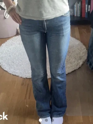 Blå bootcut jeans - Säljer ett par blå bootcut jeans med lätt slitna detaljer och klassisk femficksmodell. Jeansen har normal midja och är tillverkade i denim med lite stretch för skön passform. Perfekta för en avslappnad och trendig look.