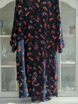 Mörkblå blommig klänning - Söt mörkblå klänning från XLNT med färgglatt blommönster i rött, orange, lila och vitt. Klänningen har lös passform, trekvartsärmar och volangdetaljer nedtill. 