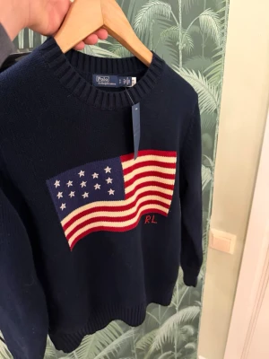 Ralph Lauren stickad tröja - Stickad mörkblå tröja från Polo Ralph Lauren med amerikansk flagga i rött, vitt och blått på bröstet samt RL-broderi. Tröjan har rund halsringning och ribbade muddar. Perfekt för dig som gillar klassisk preppy stil.