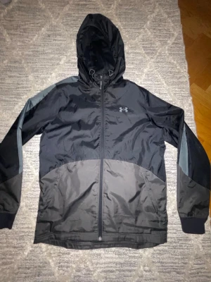 Svart vindjacka från Under Armour - Snygg svart vindjacka från Under Armour med huva och dragkedja framtill. Jackan har gråa detaljer på ärmarna och sidfickor. Tillverkad i lätt, vindtätt material som passar perfekt för blåsiga dagar. Under Armour-logga på bröstet ger en sportig look.