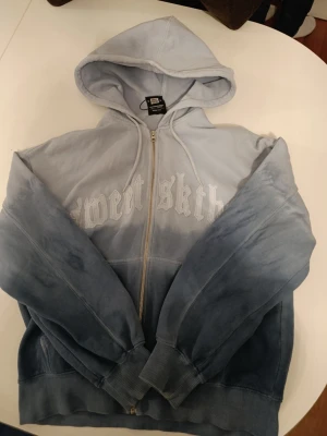 Blå hoodie från Sweet Sktbs - Säljer en blå zip-hoodie från Sweet Sktbs med snygg gradient-effekt och broderad logga på bröstet. Hoodien har huva med dragsko, två fickor fram och ribbade muddar. Perfekt för en chill streetwear-look.Använd en gång och tvättas. Litet igensytt hål på vänstra ärmen.