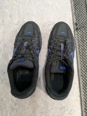 Nike svarta sneakers med blå detaljer - Säljer ett par svarta Nike sneakers med blå detaljer och meshpaneler för extra ventilation. Skorna har en sportig look med robust sula och klassisk snörning. Perfekta för dig som gillar streetwear och vill ha något stilrent men ändå med färg. Helt nya och oanvända har kvittot och boxen med.