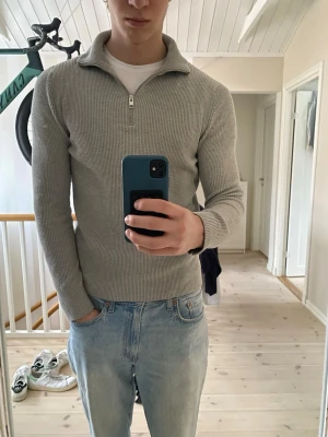 Ljusgrå ribbad half-zip tröja - Säljer en stilren ljusgrå ribbad tröja med half-zip och hög krage. Tröjan har lång ärm och är gjord i ett mjukt stickat material som passar perfekt till jeans. Enkel och clean design som funkar till många olika looks.