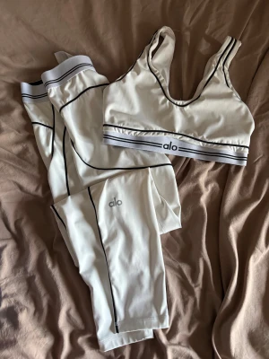 Vitt träningsset från Alo - Snyggt vitt träningsset från Alo med svarta kontrastsömmar. Setet består av en sport-bh med bred resår och matchande tights, båda med Alo-logga. Materialet är stretchigt och passar perfekt för gym eller yoga.