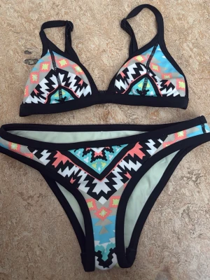 Färgglad bikini med grafiskt mönster - Snygg bikini med grafiskt mönster i svart, vit, blå, turkos, korall och gul. Överdelen har trekantskupor och svarta kanter, underdelen har matchande design. Perfekt för stranden eller poolen och ger en coolgirl look. 