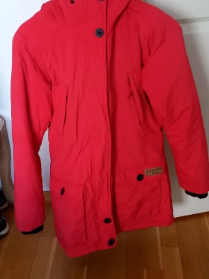 Röd parkas från North Bend - Röd parkasjacka från North Bend med huva och fuskpälsdetalj. Jackan har flera fickor med knappar framtill och dragkedja under vindslå. Perfekt för kalla dagar och har en klassisk parkasdesign.