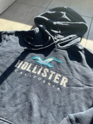 Mörkblå Hollister hoodie - Snygg mörkblå hoodie från Hollister med stor vit och turkos logga på bröstet. Klassisk känguruficka framtill och justerbar huva med dragsko. Mjuk och skön bomullsblandning, perfekt för chill dagar eller när du vill ha en avslappnad stil. Vid frågor eller funderingar skriv gärna! Passar Strl:M