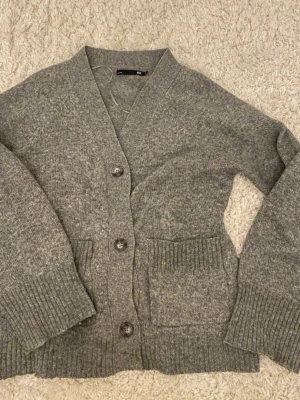 Grå kofta från lager157 med fickor - Mysig grå kofta från lager157 med v-ringning, stora knappar och två framfickor. Koftan är stickad med breda ribbade muddar vid ärmslut och nederkant. Perfekt att slänga på sig när det är kyligt ute.