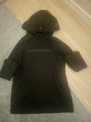 Svart hoodie från Peak Performance - Snygg svart hoodie från Peak Performance med diskret logga på bröstet. Klassisk modell med huva och långa ärmar. Tillverkad i mjukt material som känns skönt mot huden. Perfekt för en avslappnad och sportig stil.