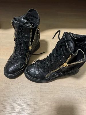 Svarta boots med krokopräglat mönster - Säljer ett par svarta stövletter med krokopräglat mönster och guldfärgade dragkedjor. Skorna har snörning framtill och detaljer i guld, vilket ger en edgy och trendig look. Tillverkade i konstläder med högre skaft och robust sula.