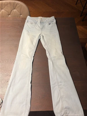 Ljusblå lågmidjande bootcut  jeans  - Säljer ett par ljusblå bootcut jeans med klassiska fem fickor och utsvängda ben. Jeansen har en hög midja och är tillverkade i ett mjukt denimtyg. Perfekta för en avslappnad och trendig look.