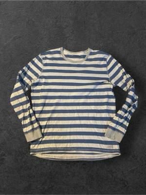 Randig långärmad tröja Peak Performance | XL - Säljer en långärmad tröja från Peak Performance med blå och vita horisontella ränder. Tröjan har rund halsringning och ribbade muddar vid ärmsluten. Tillverkad i mjukt bomullsmaterial, perfekt för en avslappnad stil.