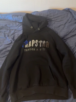Svart Trapstar hoodie med tryck - Säljer en svart hoodie från Trapstar med stor huva och magficka. Framtill står det 'Trapstar' i blått, vitt och grått samt texten 'It's a secret' i vitt. Hoodien har ribbade muddar och är gjord i mjukt bomullsmaterial.
