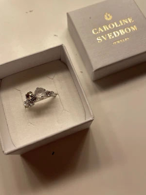 Caroline Svedbom silverring med stenar - Snygg silverfärgad ring från Caroline Svedbom med flera gnistrande stenar i olika storlekar och färger, bland annat klar och champagnefärgad. Ringen har en modern och elegant design som verkligen sticker ut. Säljer pga den är för liten