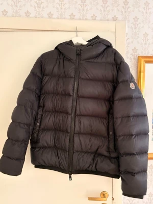 Svart dunjacka från Moncler: MYOSOTIS GIUBBOTTO - Säljer en exklusiv Moncler MYOSOTIS GIUBBOTTO i storlek 5 – använd max två gånger och i absolut nyskick.  Det här är en stilren, svart dunjacka med perfekt passform och lyxig känsla rakt igenom. Den är varm utan att kännas klumpig och passar lika bra till vardag som till en mer uppklädd look. Klassisk Moncler-logga på ärmen, kraftig tvåvägsdragkedja och en snygg, minimalistisk design som aldrig går ur tiden.