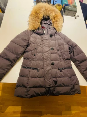 Moncler dunjacka med pälshuva - Snygg mörkgrå dunjacka från Moncler med stor fluffig pälskant på huvan. Jackan har dubbelknäppning, två dragkedjefickor fram och klassisk Moncler-logga på ärmen. Perfekt för kalla vinterdagar och riktigt varm tack vare dunfyllningen. Nypris: 13000kr