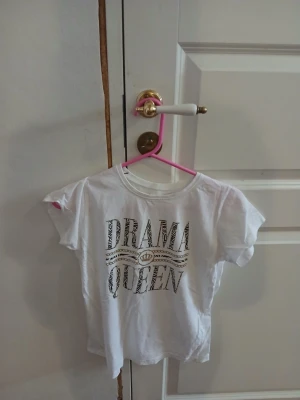 Vit t-shirt med DRAMA QUEEN-tryck - Vit t-shirt i bomull med texten DRAMA QUEEN i stort tryck framtill. T-shirten har rund halsringning och korta ärmar. Trycket har en lätt glittrig effekt och coolt mönster i bokstäverna. Perfekt för dig som vill sticka ut lite extra.