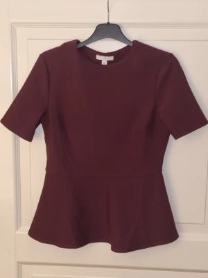 Peplumtopp vinröd  - En kortärmad topp i crêppad trikå med rund halsringning, insnitt vid bysten och utställt skört.