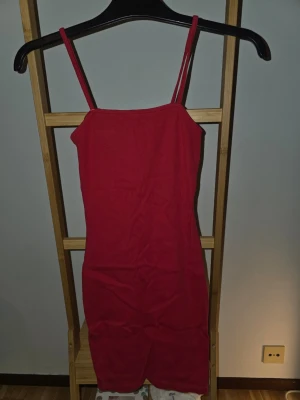 Röd tight kortklänning med smala band - Säljer en röd, tight kortklänning med smala axelband. Klänningen har en enkel, rak design och är perfekt för dig som gillar minimalistisk stil. Materialet känns stretchigt och bekvämt, vilket gör den lätt att bära hela dagen.