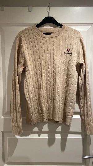 Beige kabelstickad tröja Bondelid - Snygg beige kabelstickad tröja från Bondelid med rund halsringning och broderad logga på bröstet. Tröjan har ribbade muddar vid ärmslut och nederkant. Perfekt för lager på lager och enkel att matcha med jeans. Pris går att diskutera